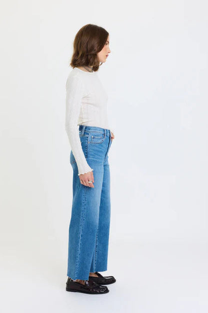 A woman shown in profile wearing a white ribbed long-sleeve top, high-waisted wide-leg blue jeans, and dark loafers against a plain white studio background.