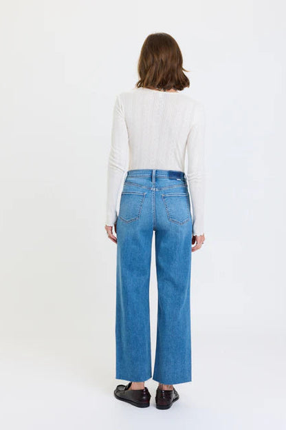 A model shown from the back wearing a white ribbed long-sleeve top, high-waisted wide-leg blue jeans, and dark brown loafers, set against a plain white studio background.