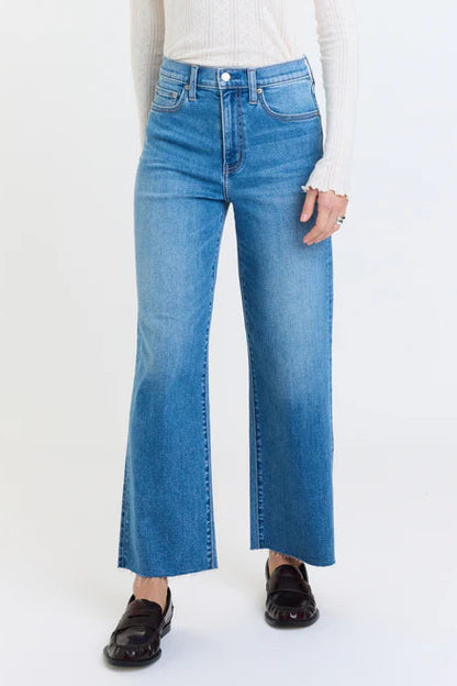 A person wearing medium-wash blue wide-leg cropped jeans with a white ribbed long-sleeve top and dark brown loafers, shown against a plain white studio background.