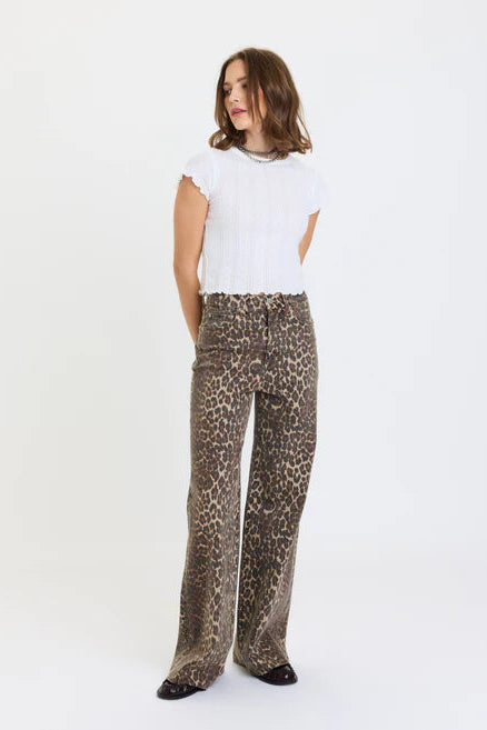 A woman modeling a pair of leopard-print wide-leg trousers with a white ribbed short-sleeve top, standing against a plain white studio background.