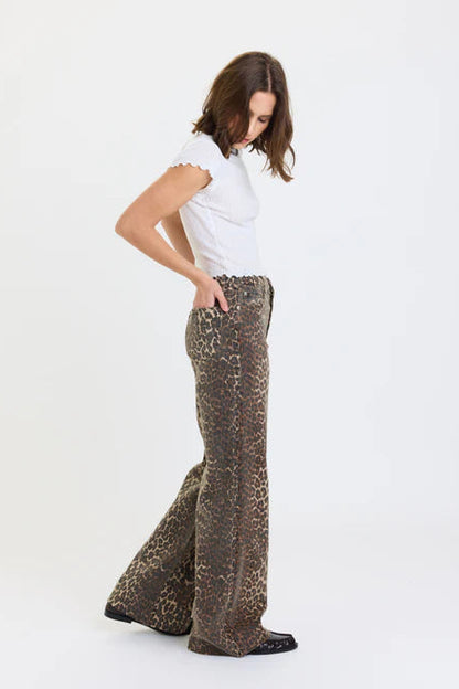 A woman modeling a pair of brown and beige leopard-print flared pants with a white ruffled short-sleeve top, standing against a plain white studio background.