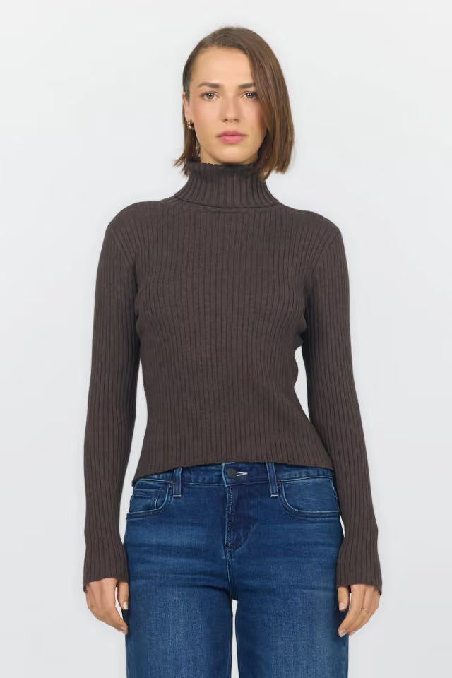 A woman modeling a dark brown ribbed knit mock neck sweater with blue jeans, shot against a plain light gray background.