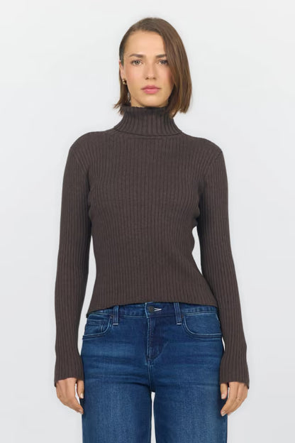 A woman modeling a dark brown ribbed knit mock neck sweater with blue jeans, shot against a plain light gray background.