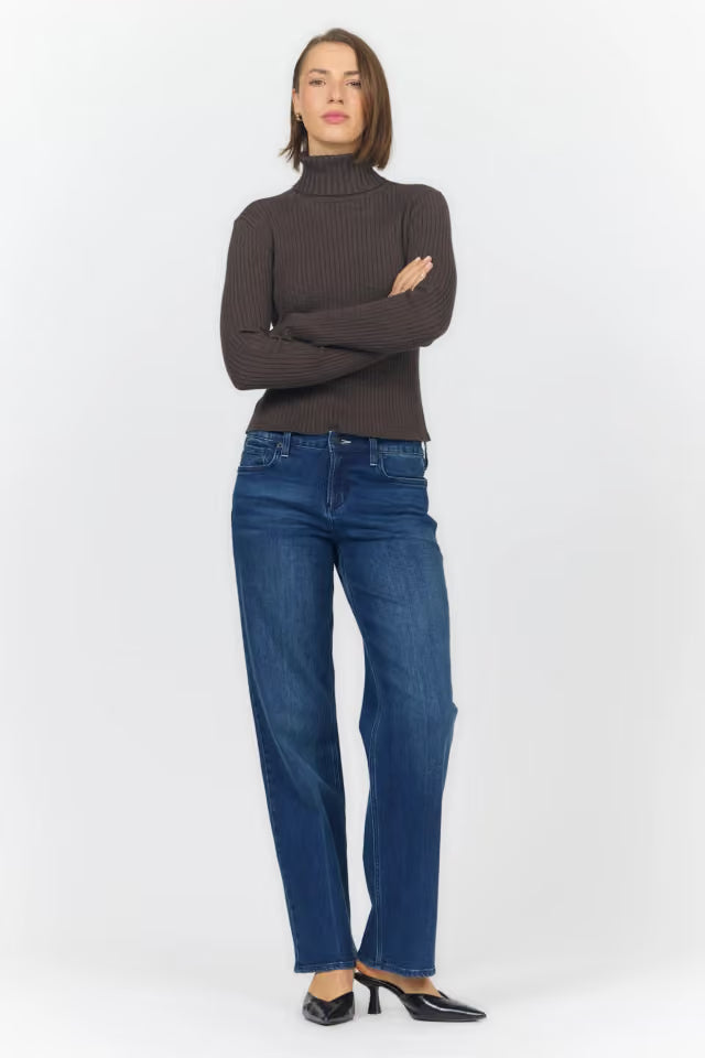 A woman modeling a dark brown ribbed turtleneck sweater and blue straight-leg jeans with black pointed-toe heels, standing against a light gray studio background.