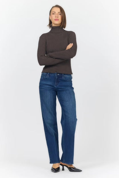 A woman modeling a dark brown ribbed turtleneck sweater and blue straight-leg jeans with black pointed-toe heels, standing against a light gray studio background.