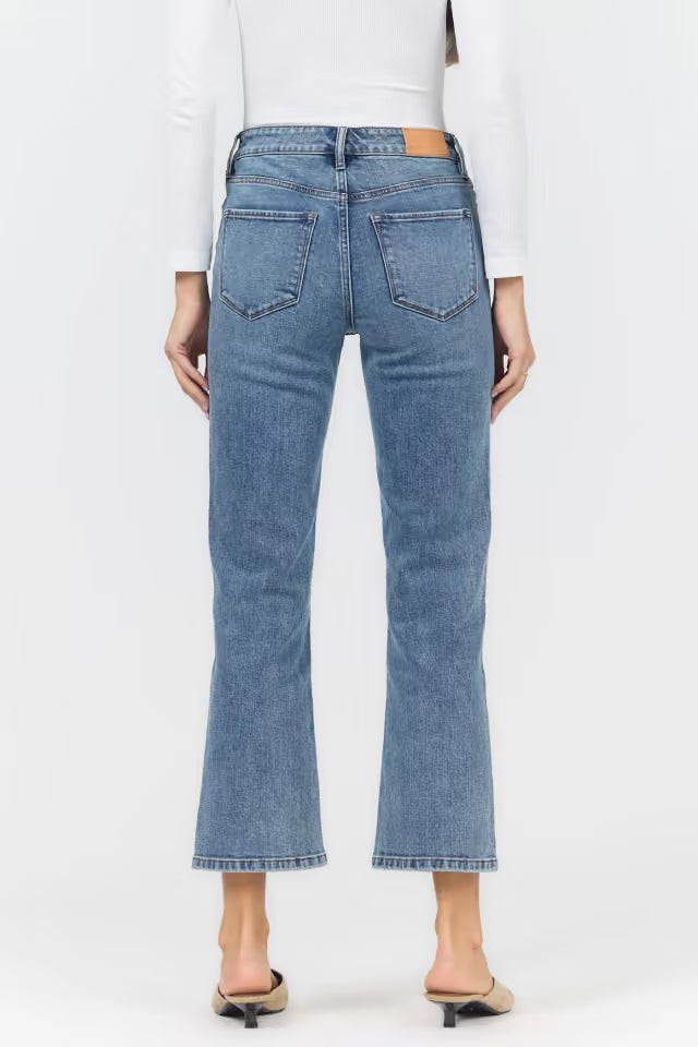 A person shown from the back wearing light blue cropped flare jeans, a white long-sleeve top, and beige pointed-toe mule heels against a plain light gray studio background.