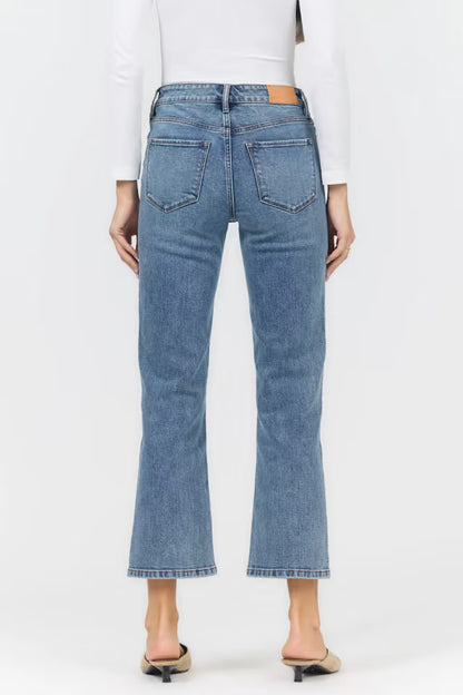 A person shown from the back wearing light blue cropped flare jeans, a white long-sleeve top, and beige pointed-toe mule heels against a plain light gray studio background.