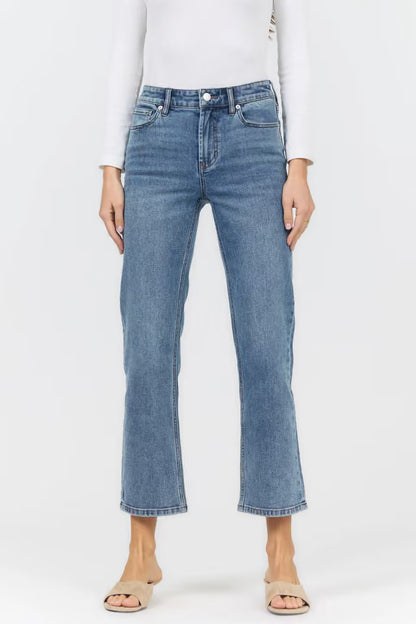A model wearing a pair of medium-wash blue cropped jeans with a straight-to-slightly-flared leg, paired with a white long-sleeve top and beige flat sandals, against a plain white studio background.