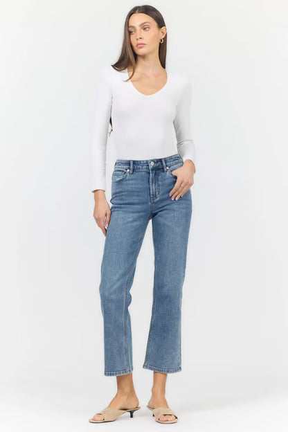 A woman modeling a pair of straight-leg blue jeans with a white long-sleeve top and beige heeled sandals, standing against a plain white studio background.