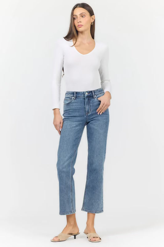 A woman modeling a pair of straight-leg blue jeans with a white long-sleeve top and beige heeled sandals, standing against a plain white studio background.
