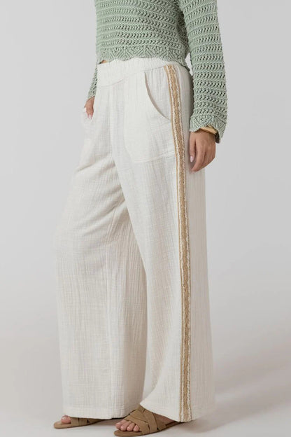 Person wearing a green knitted sweater and white pants with a gold stripe on a plain background