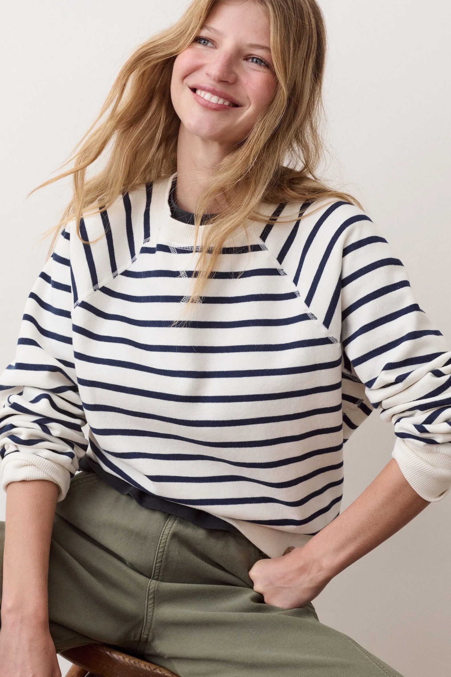 A woman wearing a cream and navy blue horizontally striped long-sleeve crewneck sweatshirt, paired with olive green pants, posing against a light beige studio background.