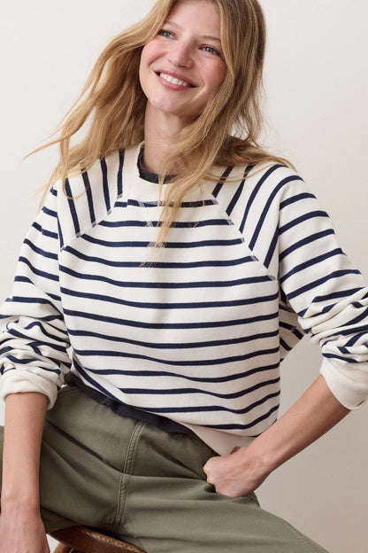 A woman wearing a cream and navy blue horizontally striped long-sleeve crewneck sweatshirt, paired with olive green pants, posing against a light beige studio background.