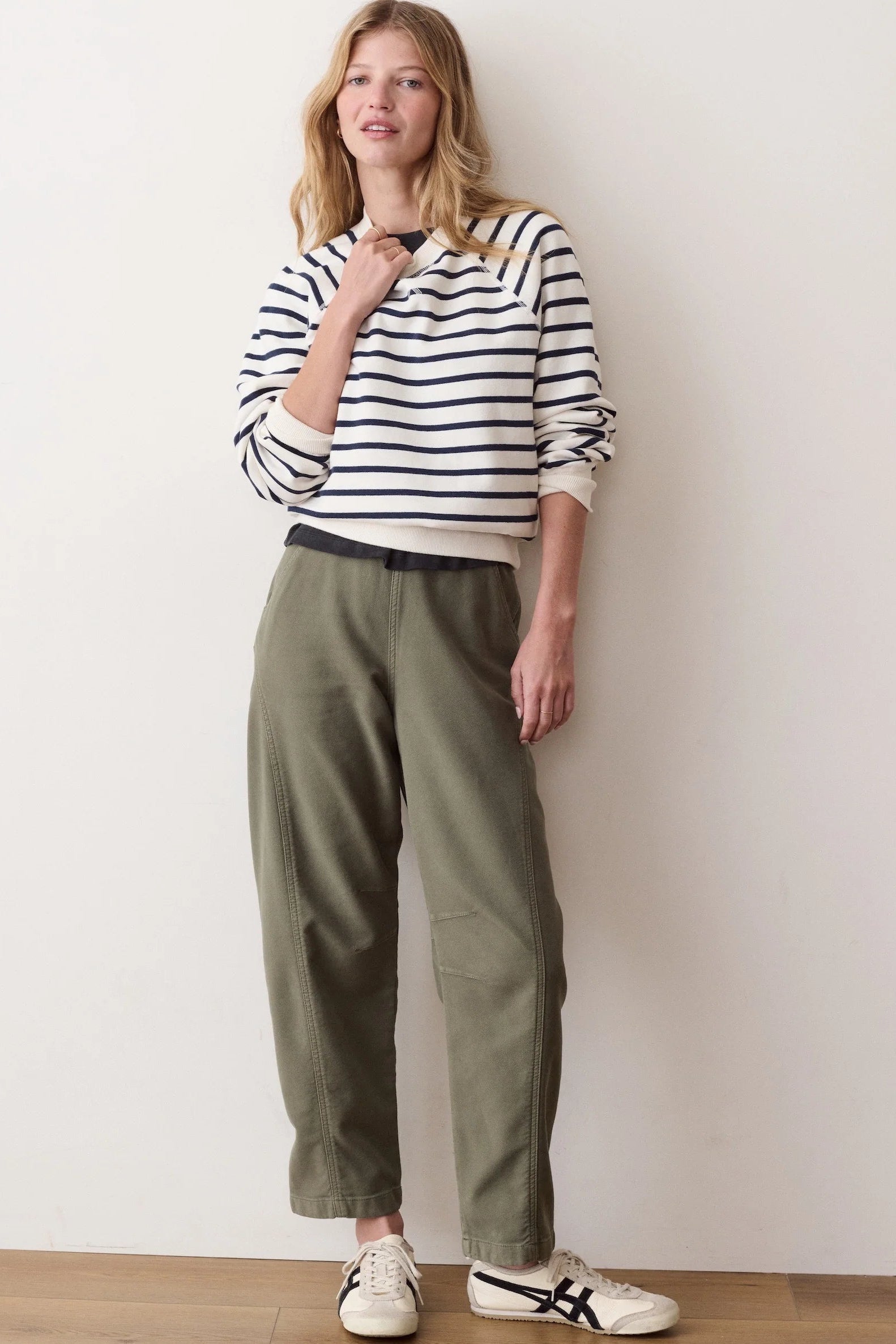 A woman stands against a plain off-white wall wearing a cream and navy blue horizontally striped long-sleeve top, olive green trousers, and white sneakers with black stripes.
