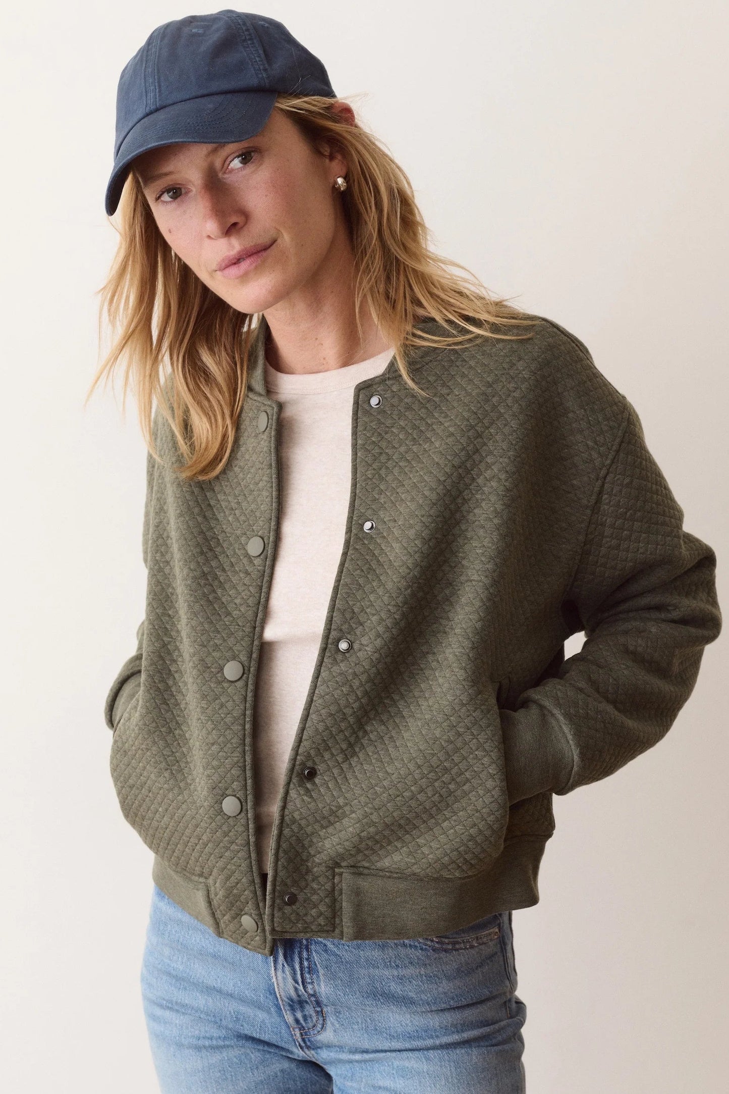 A woman wearing a textured olive green quilted cardigan over a cream t-shirt, blue jeans, and a navy blue cap, photographed against a plain light beige background.