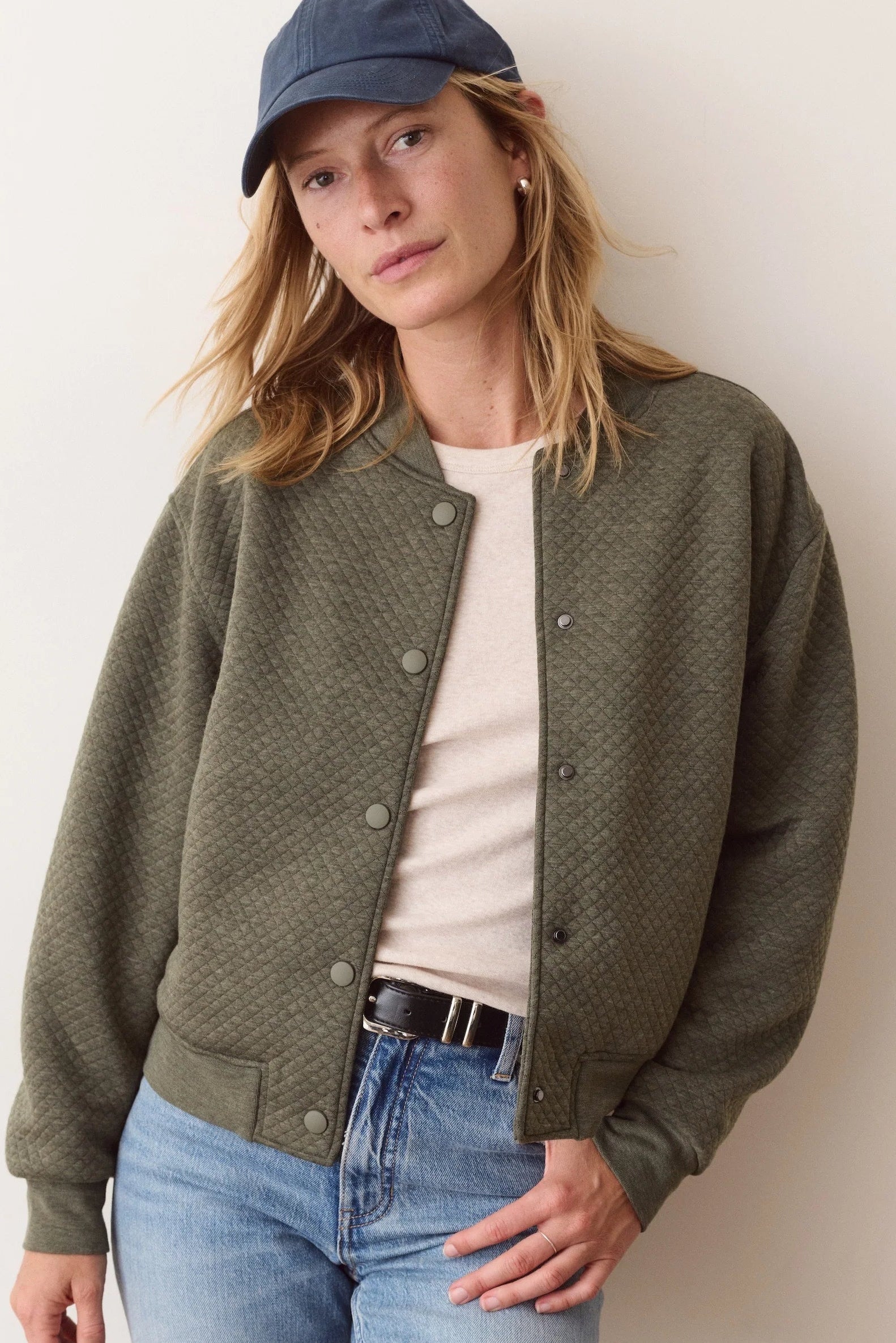 A woman wearing an olive green textured button-up cardigan over a cream t-shirt, paired with blue jeans and a navy blue baseball cap, standing against a plain off-white wall.