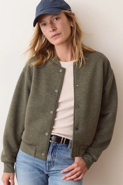 A woman wearing an olive green textured button-up cardigan over a cream t-shirt, paired with blue jeans and a navy blue baseball cap, standing against a plain off-white wall.