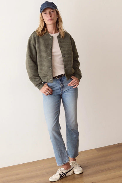 A woman stands in a minimalist room wearing an olive green knit cardigan over a cream t-shirt, light-wash blue jeans, white sneakers, and a navy blue baseball cap.
