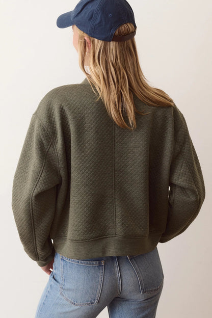 A woman shown from behind wearing an olive green textured cropped sweatshirt, blue jeans, and a navy baseball cap against a plain off-white background.