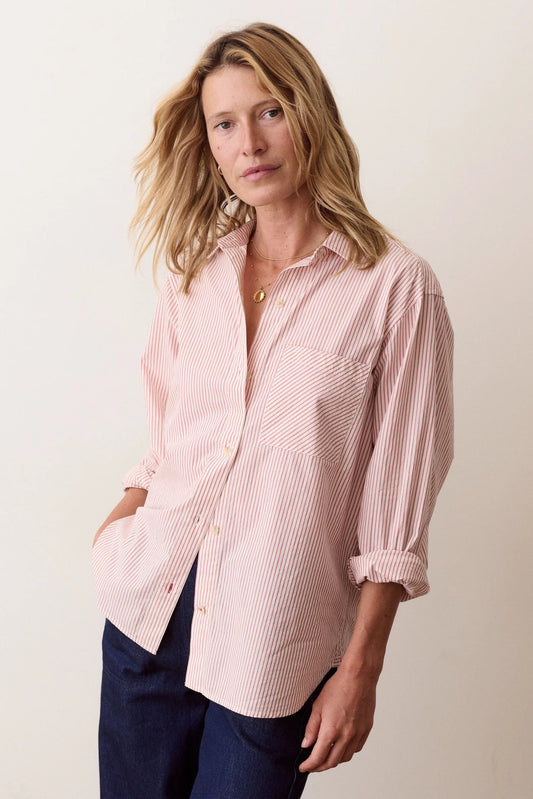 A woman wearing a light pink and white vertically striped button-down shirt with rolled-up sleeves, paired with dark blue pants, standing against a plain off-white background.
