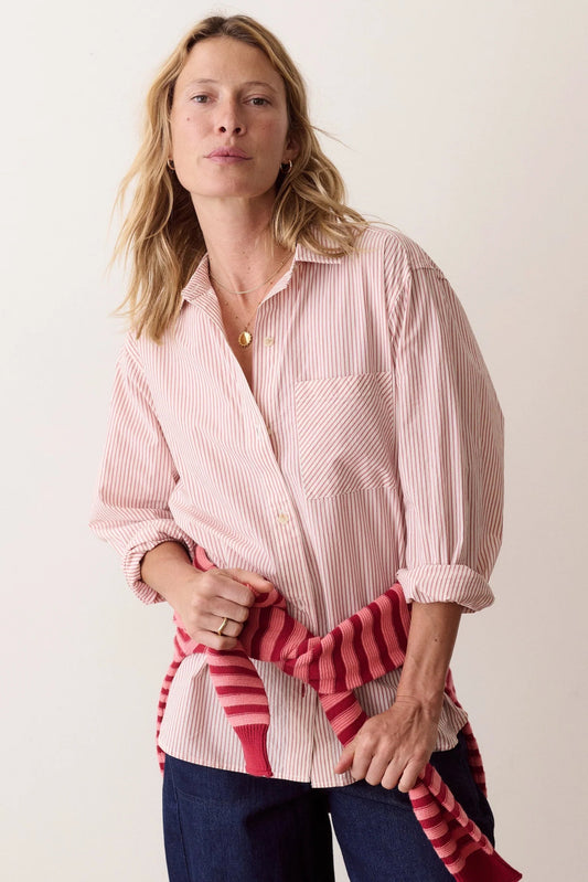A woman wearing a light pink and white vertically striped button-down shirt, with a red and pink striped knit sweater tied around her waist, paired with dark blue jeans, against a plain off-white background.