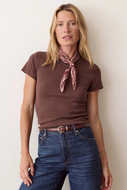A woman modeling a short-sleeve brown t-shirt with a patterned pink and white neck scarf, paired with blue jeans and a brown belt, standing against a plain off-white wall.