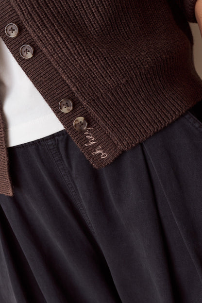 Close-up of a person wearing a dark brown ribbed knit cardigan with buttons over a white top and black skirt. The cardigan has 'stay go' embroidered in white script near the hem.
