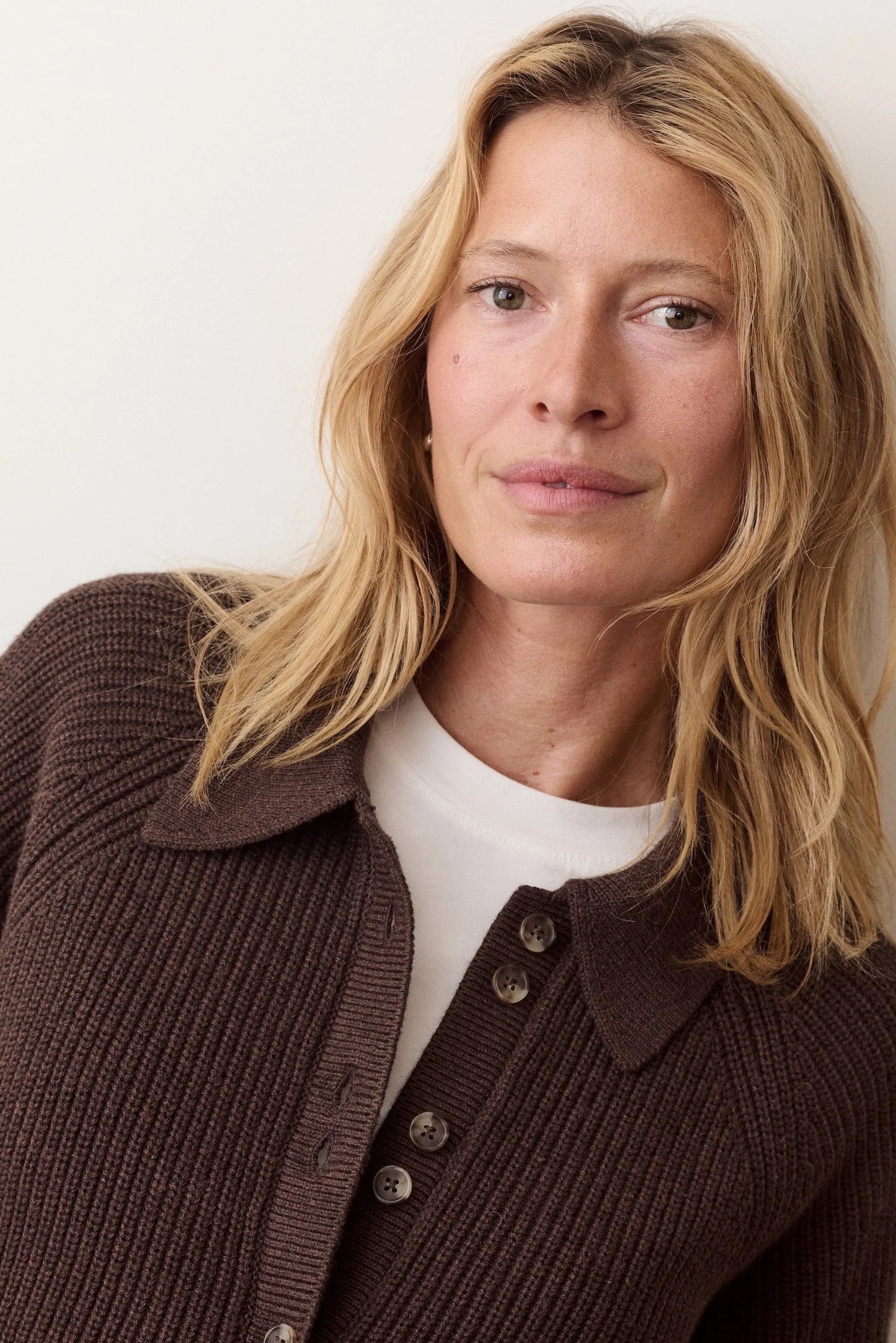 A woman with shoulder-length blonde hair wearing a dark brown ribbed knit button-front cardigan over a white crew-neck top, photographed against a plain off-white wall.