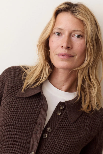A woman with shoulder-length blonde hair wearing a dark brown ribbed knit button-front cardigan over a white crew-neck top, photographed against a plain off-white wall.