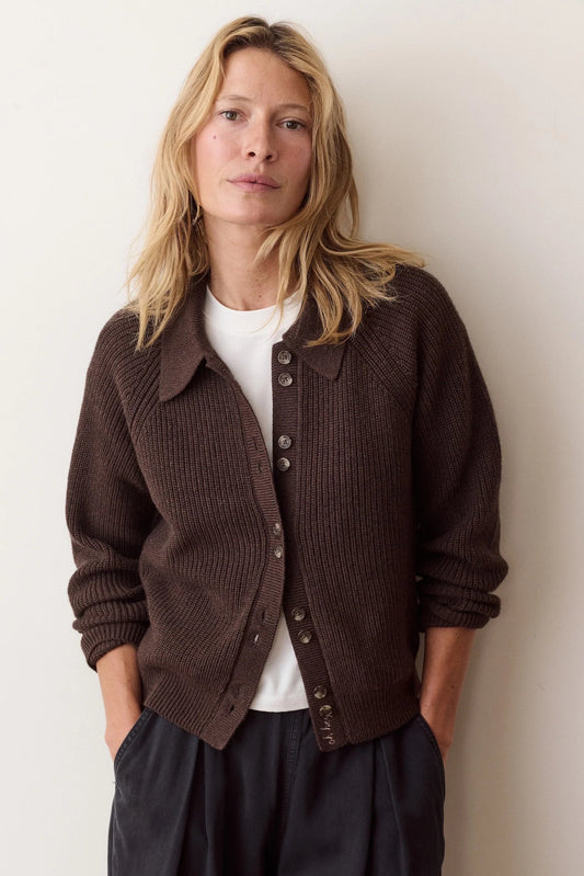 A woman with blonde hair wearing a dark brown ribbed knit cardigan over a white t-shirt and dark trousers, standing against a plain off-white wall.