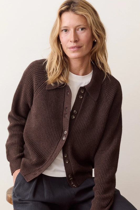 A woman wearing a dark brown ribbed knit button-front cardigan over a white top and dark trousers, seated against a plain light beige background.
