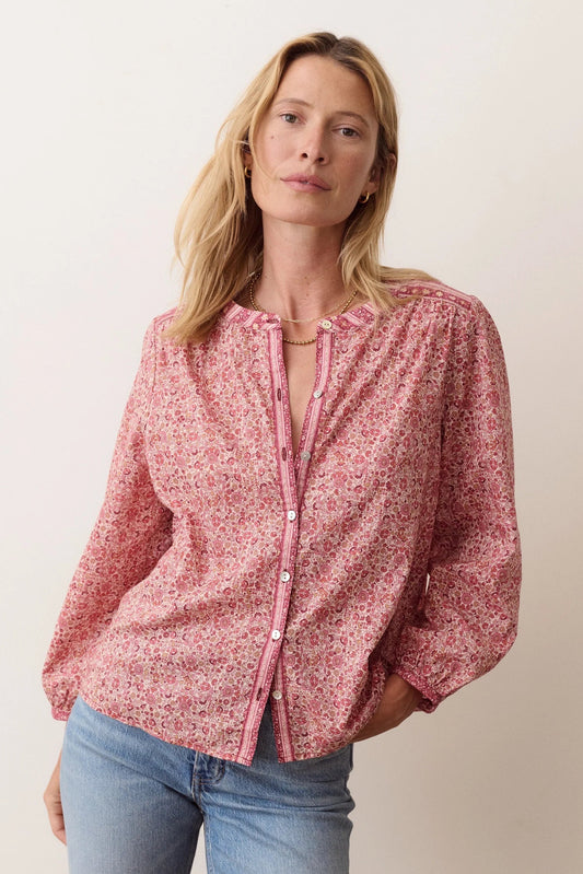 A woman wearing a pink and red floral print button-up blouse with blue jeans, standing against a plain light beige background.