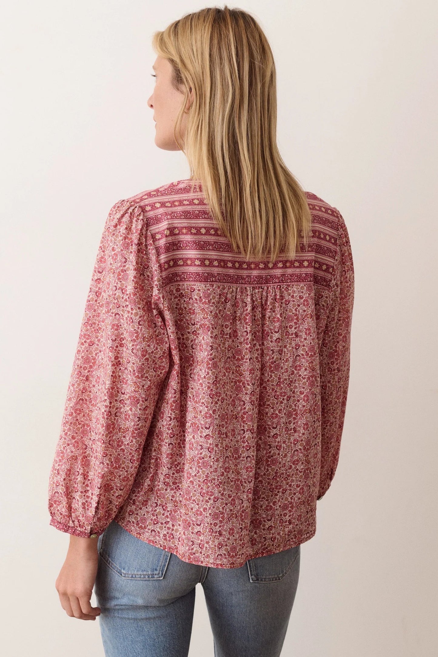 A woman shown from the back wearing a dusty rose long-sleeve blouse with a dense small floral print and horizontal embroidered detailing across the upper back, paired with blue jeans against a plain off-white background.