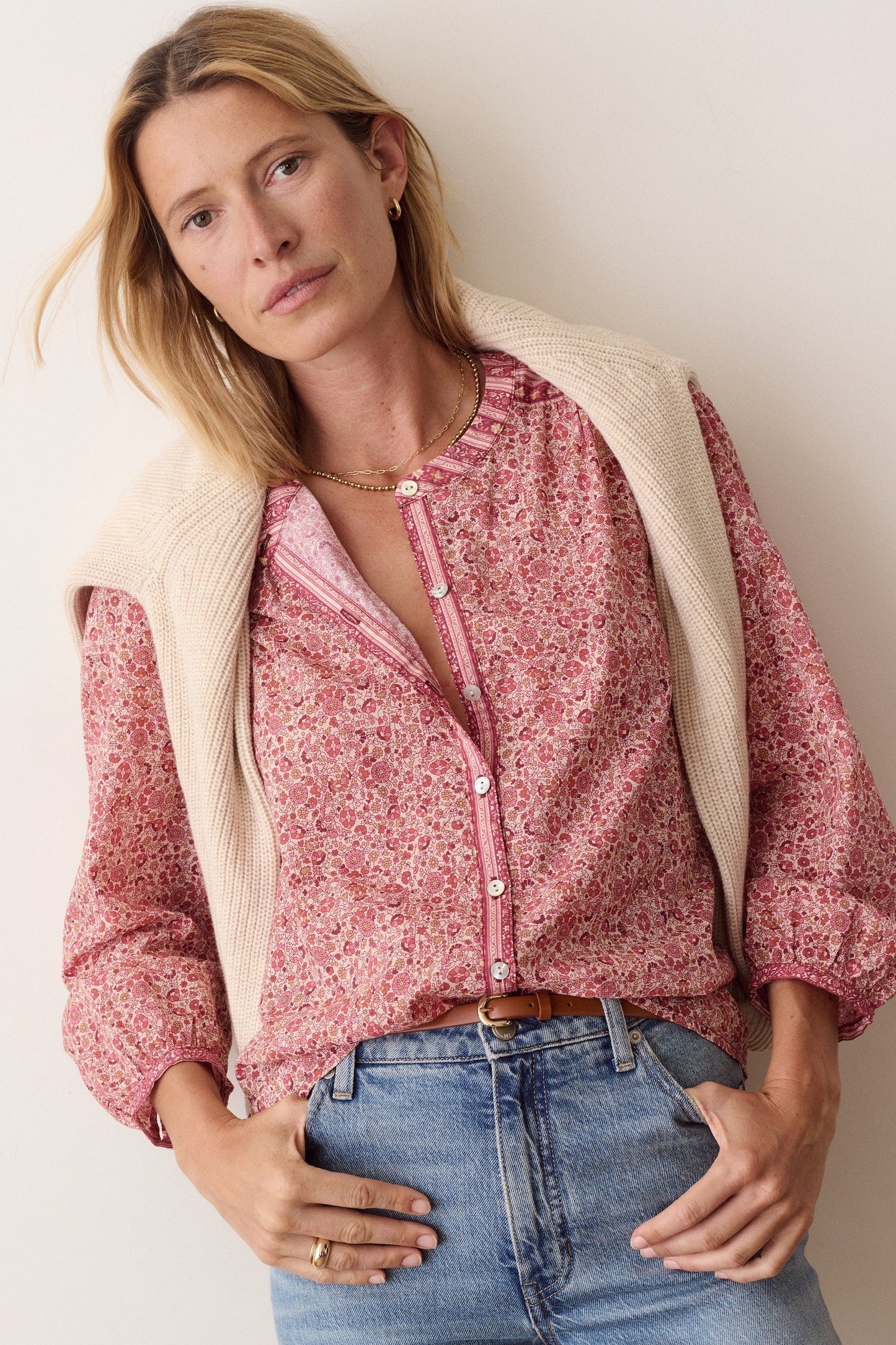 A woman wearing a pink floral long-sleeve button-up shirt with a cream ribbed knit vest and blue jeans, posing against a plain light beige wall.