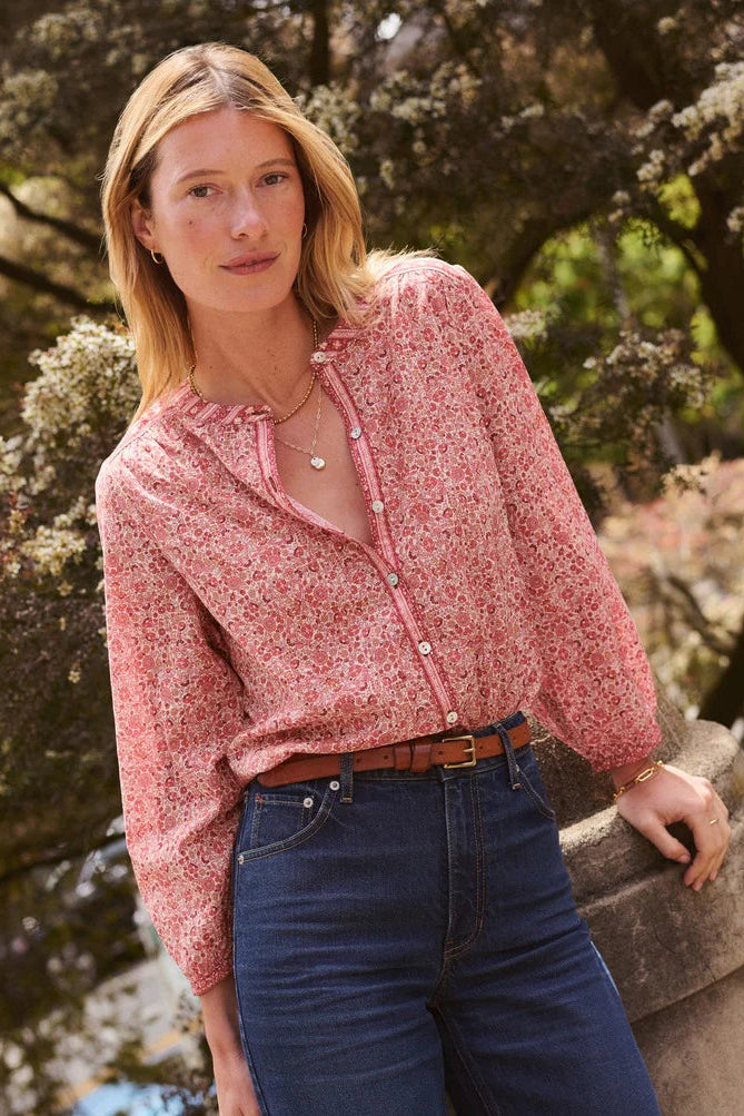 A woman wearing a pink and white floral long-sleeve button-up blouse tucked into dark blue jeans with a brown belt, standing outdoors against a background of trees and foliage.