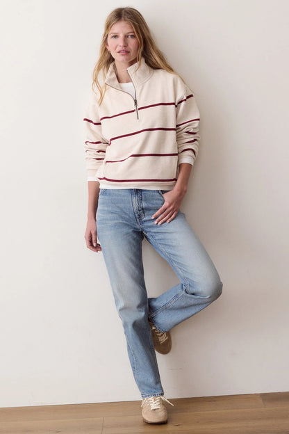 A woman stands against a plain off-white wall wearing a cream quarter-zip pullover sweater with burgundy stripes, light-wash blue jeans, and beige sneakers.