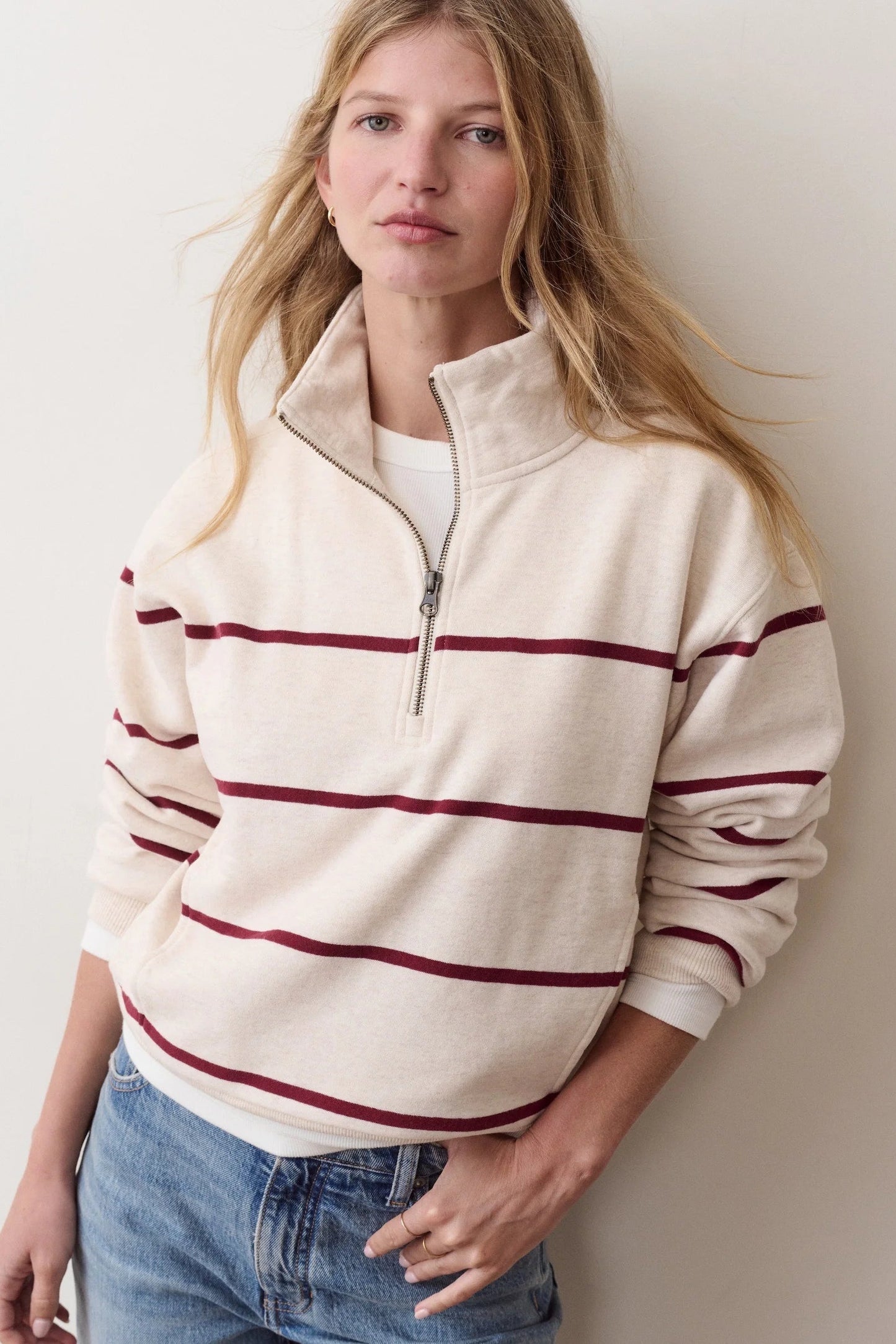 A woman wearing a cream-colored quarter-zip sweatshirt with dark red horizontal stripes, paired with blue jeans, standing against a plain off-white wall.