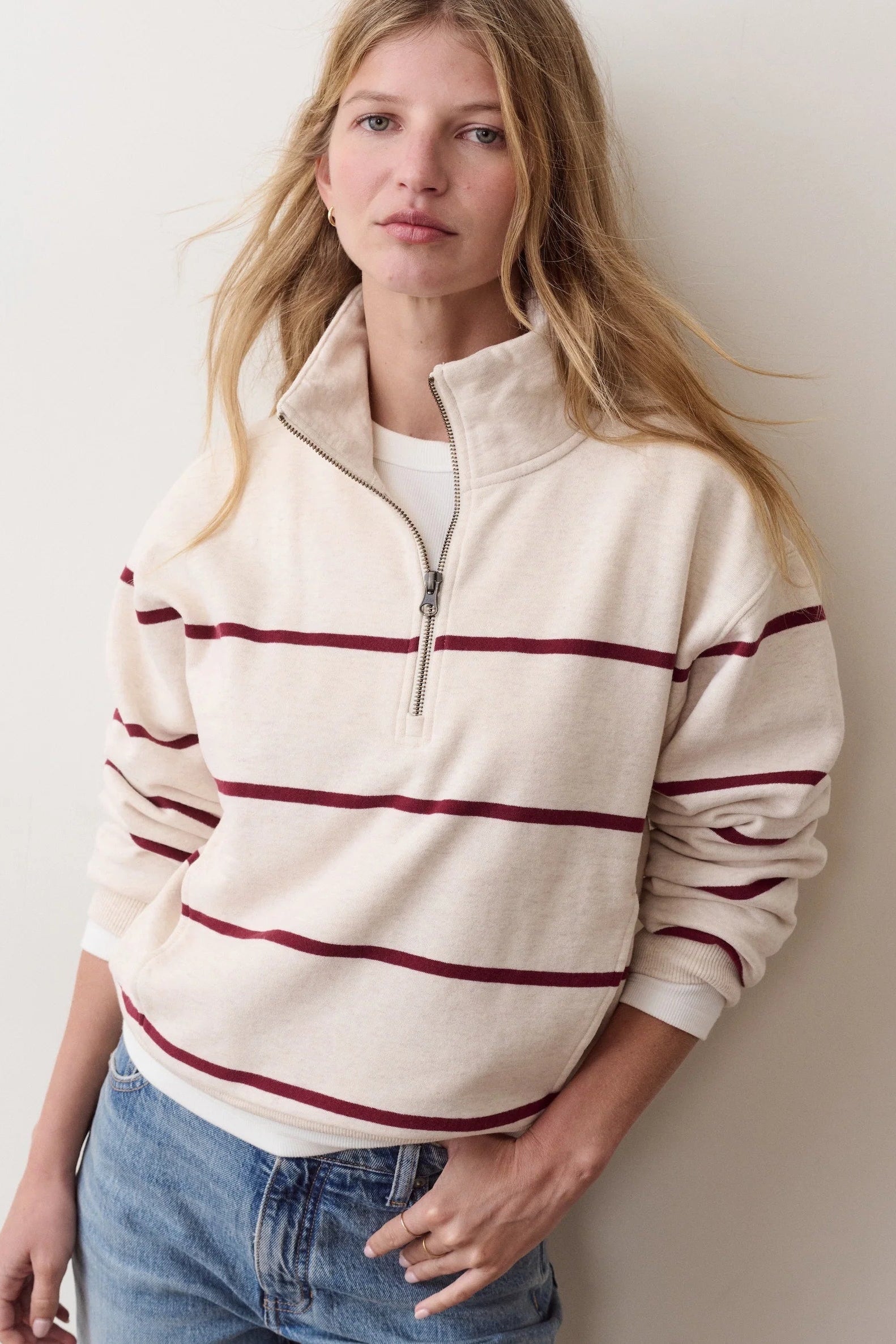 A woman wearing a cream-colored quarter-zip sweatshirt with dark red horizontal stripes, paired with blue jeans, standing against a plain off-white wall.