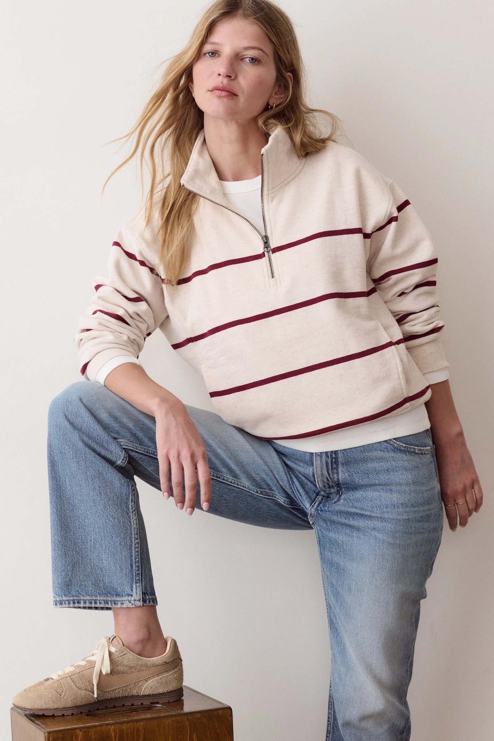 A woman models a cream-colored quarter-zip sweatshirt with burgundy horizontal stripes, paired with light-wash blue jeans and beige sneakers, sitting on a wooden block against a plain off-white wall.