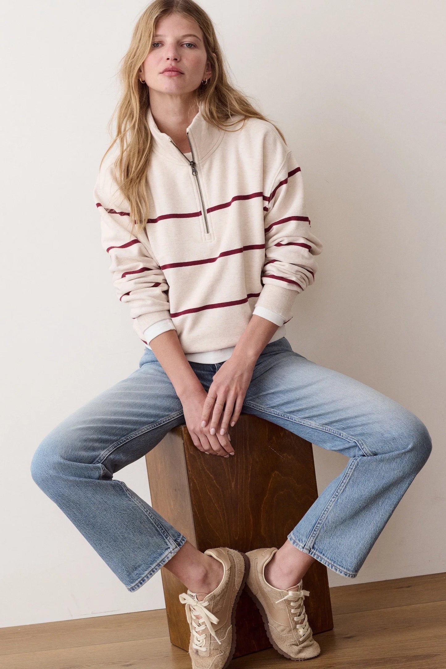 A woman sits on a wooden block wearing a cream quarter-zip pullover sweater with burgundy stripes, light-wash blue jeans, and beige sneakers against a plain off-white wall.
