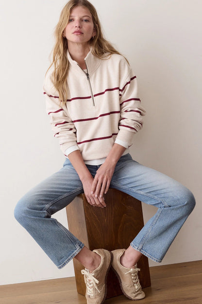 A woman sits on a wooden block wearing a cream quarter-zip pullover sweater with burgundy stripes, light-wash blue jeans, and beige sneakers against a plain off-white wall.