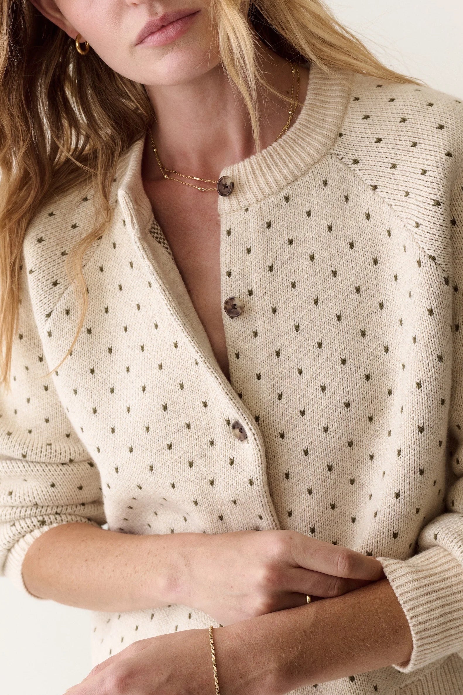 A close-up of a woman wearing a cream-colored knit cardigan with small dark brown polka dots and dark brown buttons, styled with delicate gold jewelry against a clean off-white background.