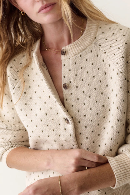 A close-up of a woman wearing a cream-colored knit cardigan with small dark brown polka dots and dark brown buttons, styled with delicate gold jewelry against a clean off-white background.