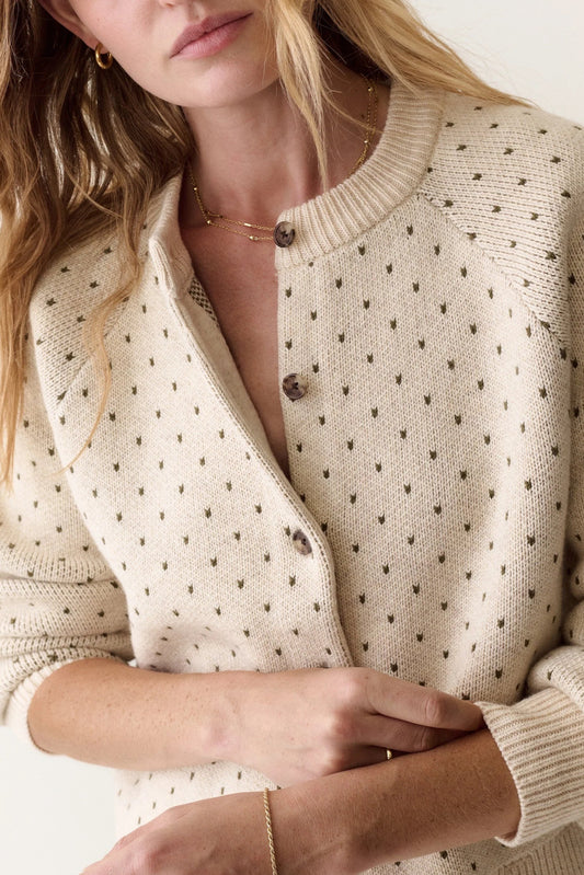 A close-up of a woman wearing a cream-colored knit cardigan with small dark brown polka dots and dark brown buttons, styled with delicate gold jewelry against a clean off-white background.