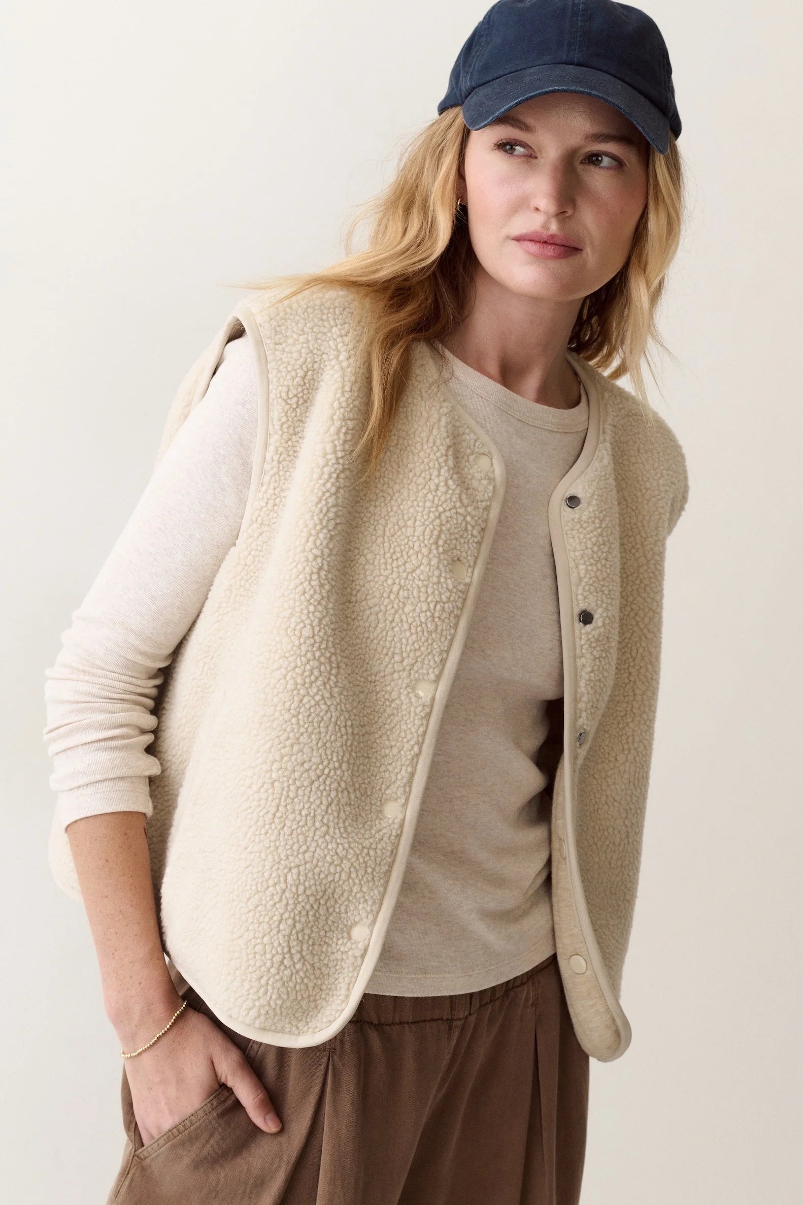 A woman wearing a beige sherpa fleece button-front vest over a cream long-sleeve top, brown pants, and a navy baseball cap, photographed against a plain light beige studio background.