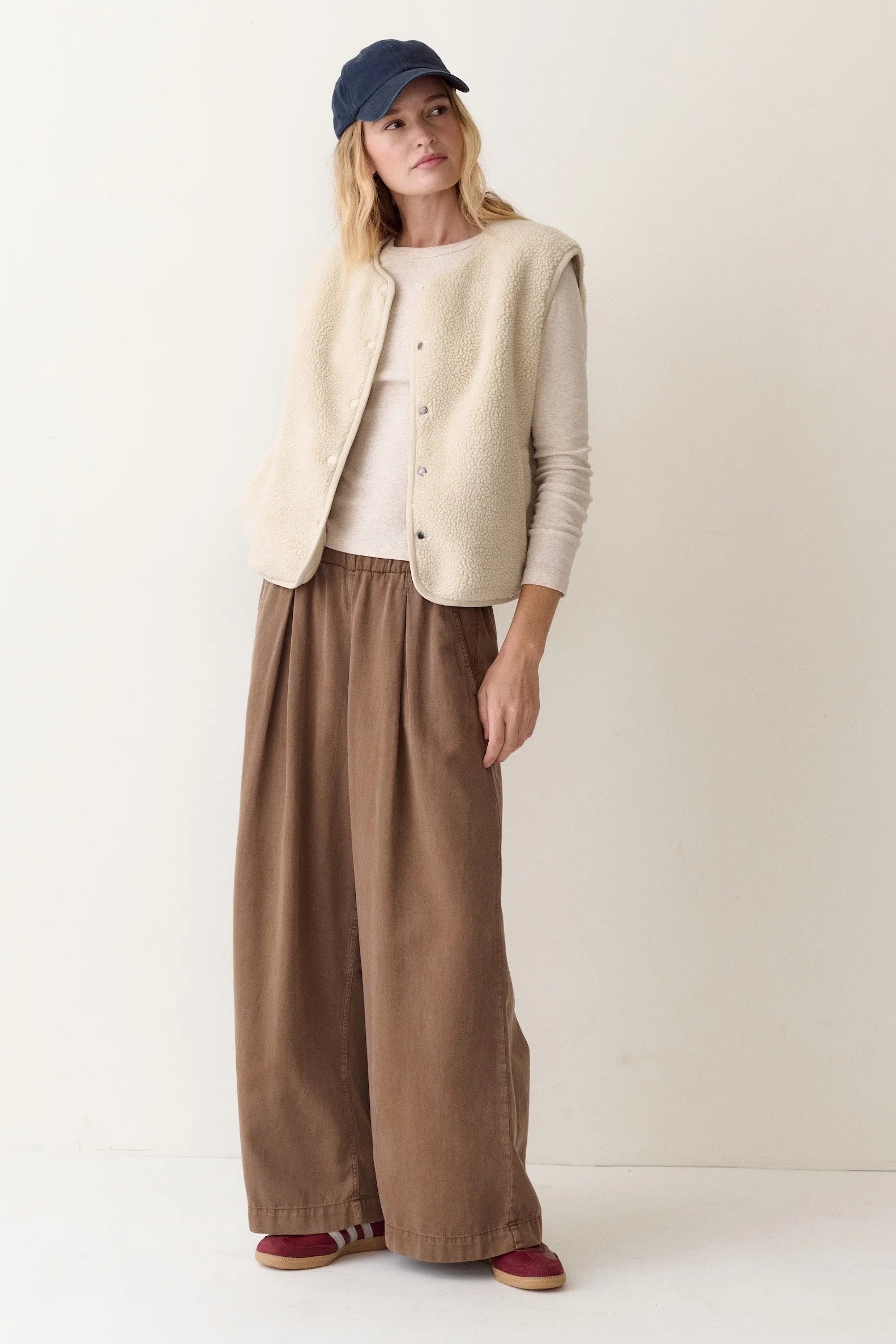 A woman stands against a plain off-white studio background wearing a cream sleeveless button-front vest over a light beige long-sleeve top, wide-leg brown pants, a navy cap, and burgundy shoes.