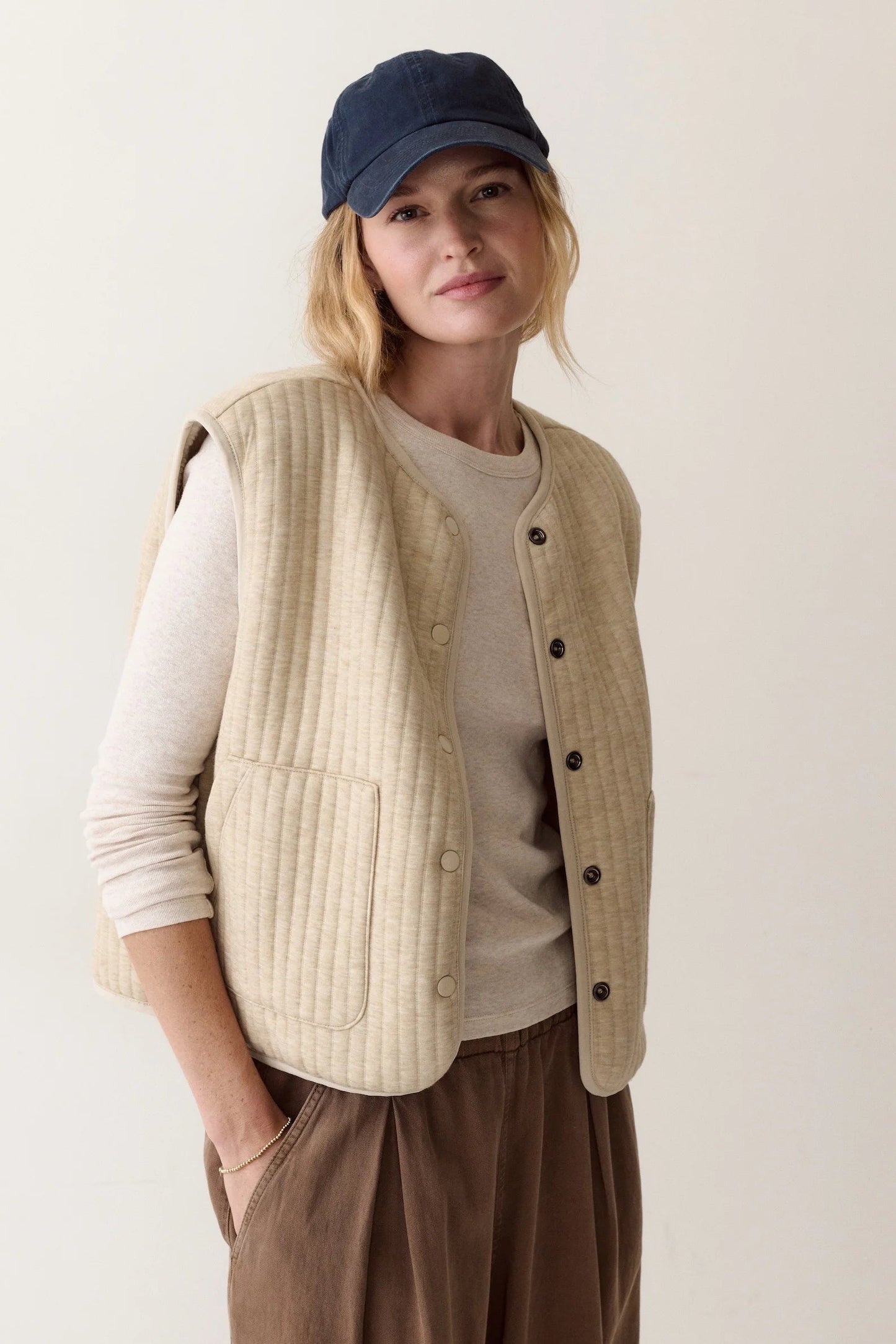 A woman wearing a beige ribbed button-front vest over a cream long-sleeve top, brown pants, and a navy baseball cap, standing against a plain light beige studio background.