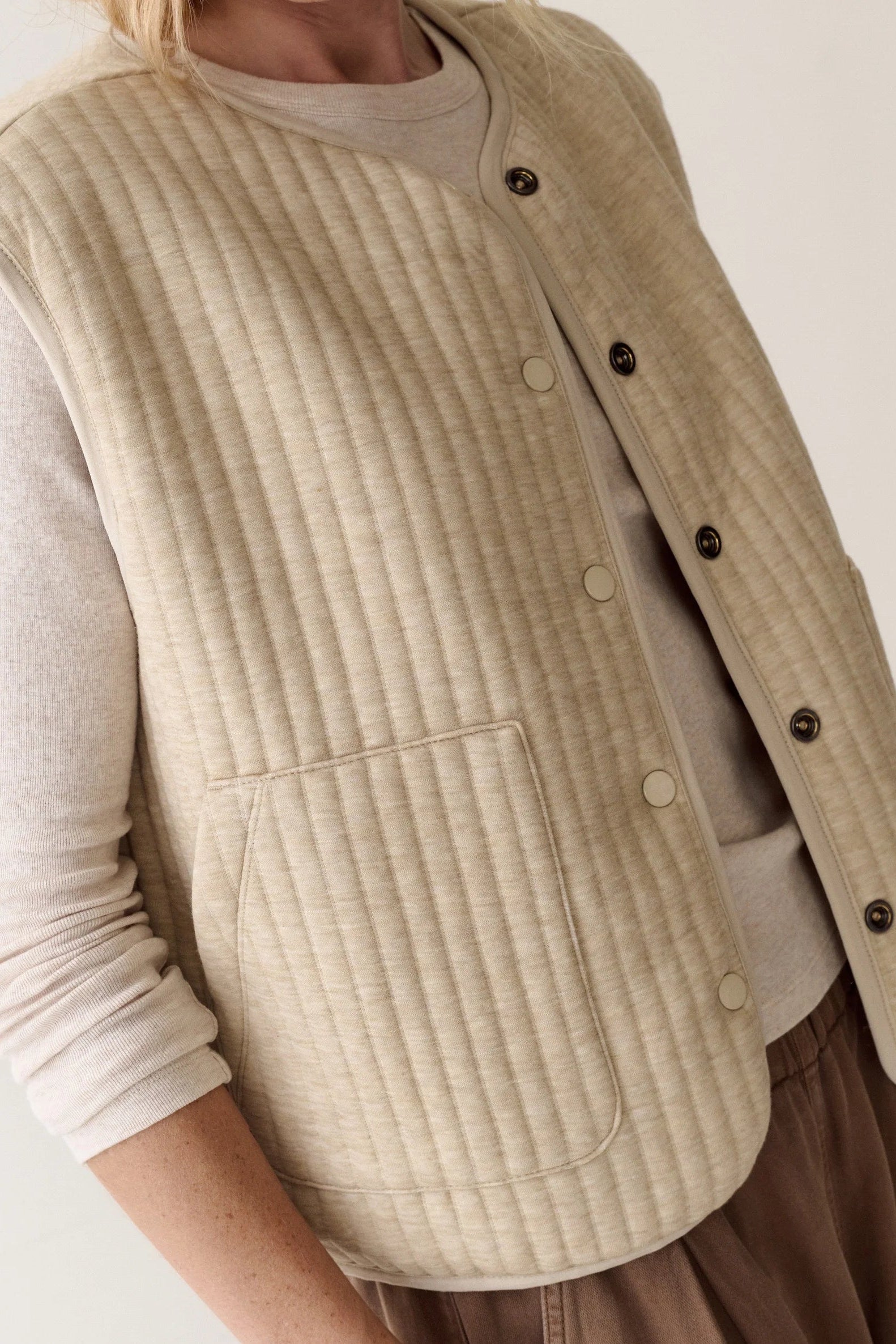 A close-up of a person wearing a sleeveless quilted vest over a light beige long-sleeve shirt. The vest is ribbed and button-front with dark buttons, and a large front patch pocket is visible.