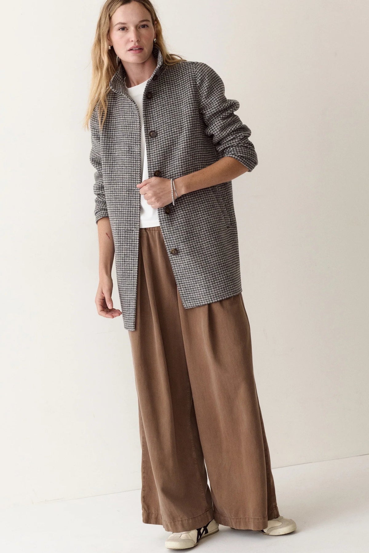 A woman modeling a gray and black patterned jacket over a white top and brown wide-leg trousers, standing against a plain off-white studio background.