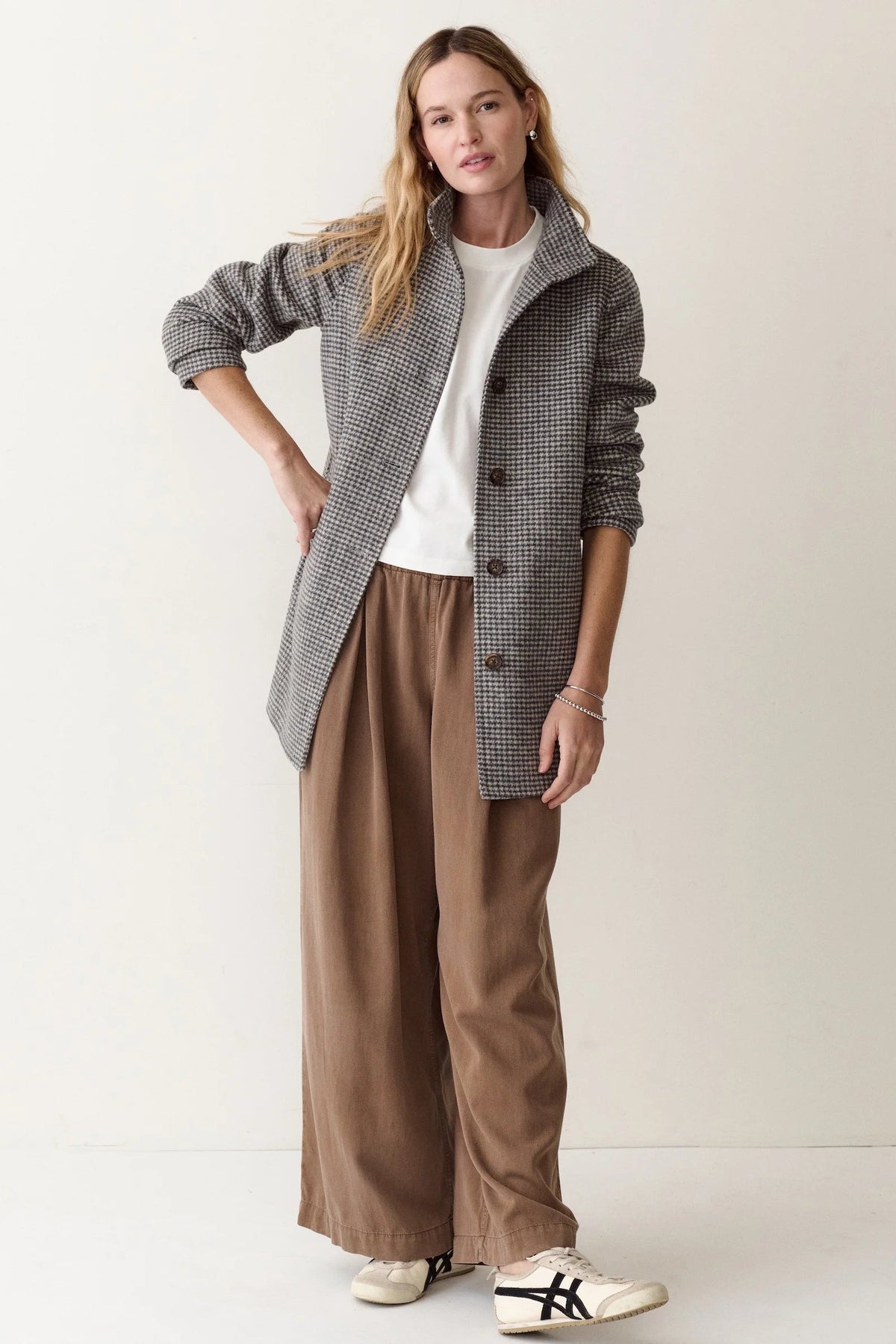 A woman modeling a stylish outfit consisting of a gray and black houndstooth button-up jacket, brown wide-leg trousers, a white t-shirt, and white sneakers with black stripes, set against a plain off-white studio background.
